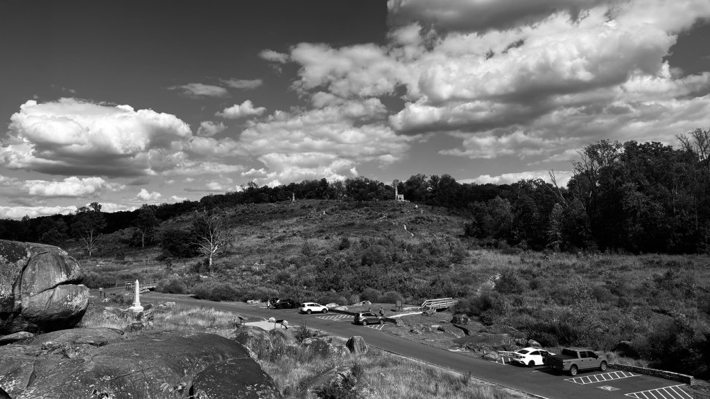Black-and-white wide photograph of Little Round Top in Gettysburg National Military Park, showing the hillside monument, large foreground boulders, parked cars below, and a dramatic cloud-filled sky.
