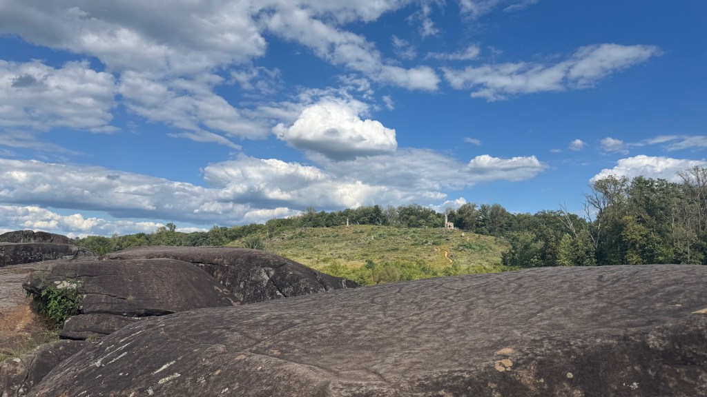 Color photograph of Little Round Top in Gettysburg National Military Park, taken from atop large rock formations in the foreground, with green hillside, scattered monuments, and a bright blue sky filled with white clouds.