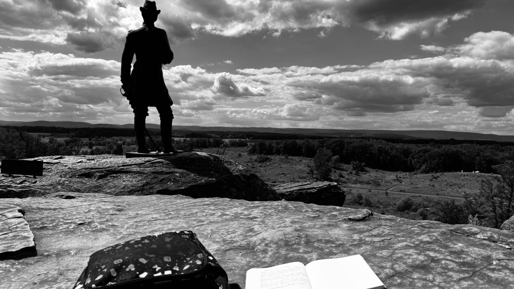Black-and-white photograph taken atop Little Round Top in Gettysburg National Military Park. In the foreground, an open notebook rests on the rock beside a patterned case, with handwritten lines visible. A silhouetted statue stands overlooking the battlefield beneath a dramatic clouded sky.