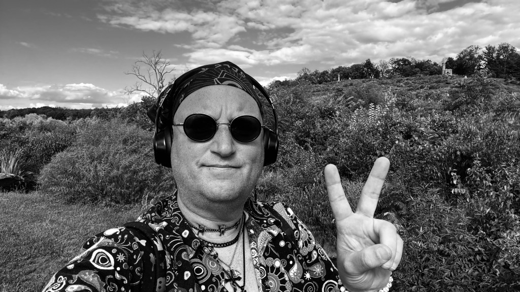 Black-and-white selfie of Tristan Robert Lange at Little Round Top in Gettysburg National Military Park, wearing round sunglasses, headphones, and a patterned shirt, holding up a peace sign with the battlefield hillside and monument visible behind him under a clouded sky.