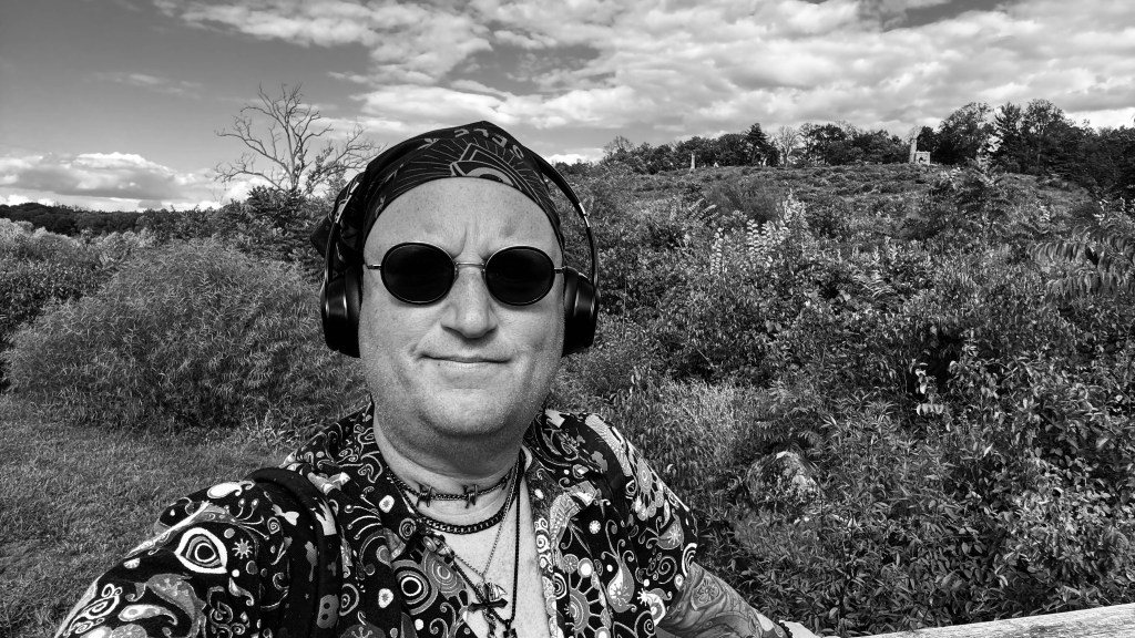 Black-and-white selfie of Tristan Robert Lange at Little Round Top in Gettysburg, wearing sunglasses, headphones, and layered necklaces, with rolling brush, trees, and a monument visible on the hill behind him beneath dramatic clouds.