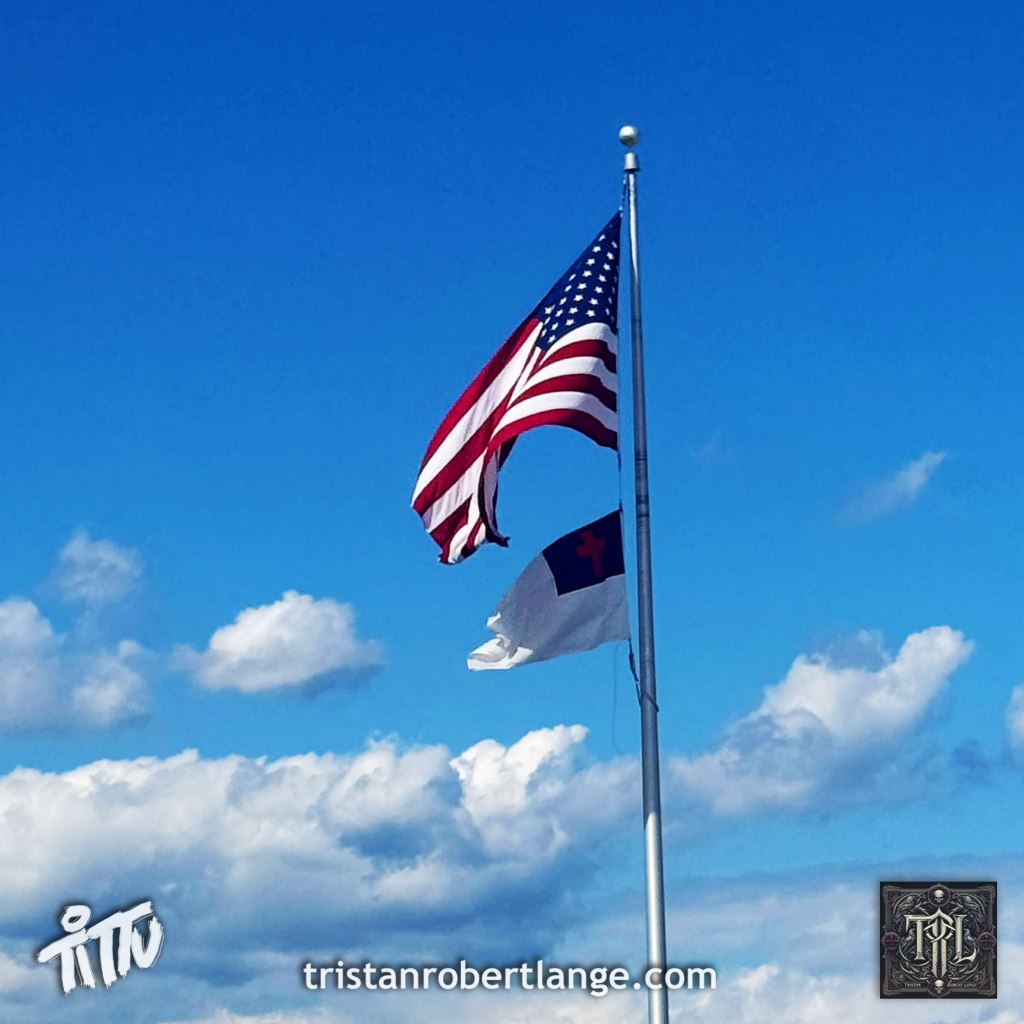 A low-angle photograph of two flags flying from the same white pole against a pale blue, lightly clouded sky. The American flag occupies the upper position, with the Christian flag flying directly beneath it.